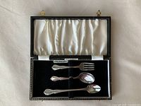 Open presentation box showing three flatware pieces on velvet insert, satin lid lining and maker label