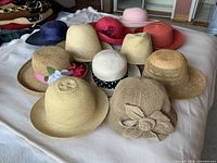 All eleven women’s straw and paper braid hats in various colours and styles arranged on a bed surface