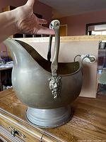 Overall view of brass coal scuttle on pedestal base with ceramic-tipped swing handle and side lion head detail