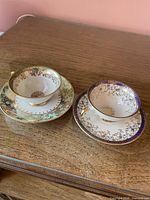 Both teacup and saucer sets side by side showing green and purple/cobalt bordered designs with gold gilt