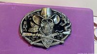 Front view of belt buckle showing skull, hat, rifles, snake design