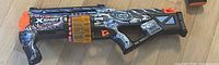 Left side view of blue ZURU X-Shot dart blaster with shark graphic, showing drum magazine and front muzzle
