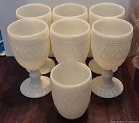 Front view of seven milk glass goblets showing embossed pattern and pedestal bases