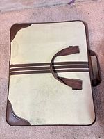 Closed beige canvas portfolio case showing brown vinyl handles, stripes and corner protectors with visible stains