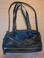 Front view of black textured handbag showing double handles, detachable strap, front zipper pockets, lower zipper compartment