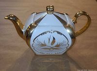 Side view of cream ceramic teapot with gold spout, handle, lid finial and floral panel