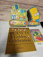 Overall lot showing two Simpsons trivia game tins, Peanuts book and Simpsons DVD