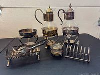 Full lot showing both glass carafes in stands, compote bowl, cracker tray, tongs, coaster stack, toast rack