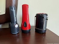 Three lighting devices: black MAXXE Workhorse flashlight, red handheld flashlight, black and red camping lantern with metal handles standing on wooden surface