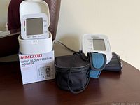 Overall view of both blood pressure monitors, wrist unit on box, arm unit with cuff and pouch