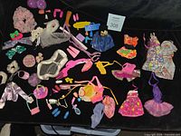 Overview of assorted doll clothes, shoes, hats, skateboard, radio, accessories on black fabric