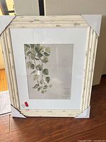 Framed wall art with distressed white frame and botanical print, protective corner guards visible