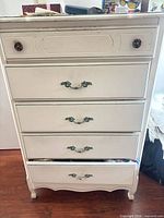 Front view of cream five-drawer dresser with ornate metal handles and scalloped base