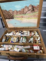 Open wooden paint box showing numerous oil paint tubes, bottles and painted panel in lid