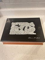 Top view showing metal relief design and artist signature on black lid