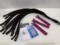 All items laid out: six long brown zippers, two short magenta zippers, Dritz button expanders pack, Dritz collar stays pack