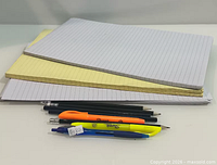 Stack of three lined writing pads with eight assorted writing instruments arranged in front