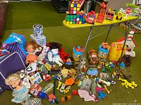 Overall view of all toys spread on carpet including dolls, plush toys, vehicles, tins, bucket, Rubik’s blocks
