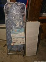 Ironing board wrapped in plastic next to two white shelves wrapped in plastic