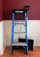 Front view of Werner 4 ft ladder standing upright, showing blue frame, three metal steps, and black tool tray with Werner logo. Artic Blast snow shovel with black plastic blade resting behind the ladder.