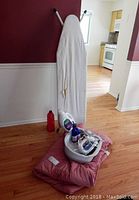 Photo shows ironing board, Sunbeam Steam Master iron, Homemedics foot massager, pink comforter, and several bottles including laundry detergent and cleaning gel.