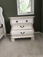 Front view of the white wooden bedside table showing three drawers and rounded feet.