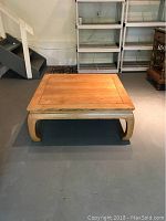 Wooden square coffee table with curved legs and paneled top placed on concrete floor with stairs in background.