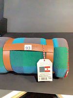 Rolled blanket secured with strap, showing plaid pattern and hang tag