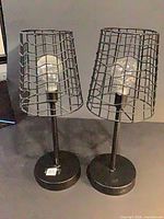 Front view of both black wire-cage table lamps with LED bulbs installed