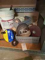 Grey crate holding several grinding wheels, sanding disc, wire brush and bucket nearby