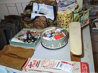 Table of sewing equipment including Janome machine in floral bag, thread tin, wooden tray, skirt marker box, mini ironing board, floral storage case