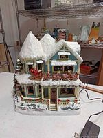 Front view of illuminated Thomas Kinkade story house showing Santa sleigh, reindeer, porch, and power cord
