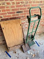 Hand truck, wooden board, and square blade shovel leaning against brick wall