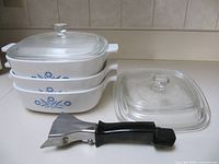 Stack of three Cornflower Blue casseroles with two lids stacked, third lid and detachable handle in foreground