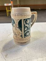 Front view of beige and green ceramic beer stein showing relief figure panel and handle
