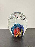 Front view showing entire oval glass paperweight with internal colorful swirl and two white cranes