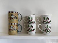Two chickadee pattern mugs stacked next to four holly pattern mugs