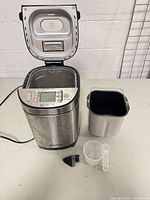 Bread maker open showing interior, bread pan, measuring cup, spoon, paddle