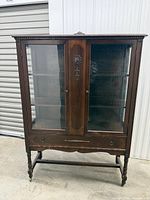Front view of wooden china cabinet showing glass doors, carved panel, lower drawer, legs and stretcher