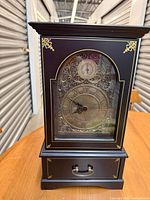 Front view of Bombay Company Tavernier Key mantel clock showing dial, glass door, gold accents, and lower drawer