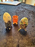 Three primary objects: porcelain egg in amethyst Fenton cup, marigold Fenton cup, Murano egg on gold stand