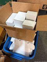 Open shipping carton holding several unfolded white BCW card boxes stacked with additional boxes in blue tote below
