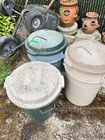 Three plastic trash cans with lids and wheelbarrow frame behind