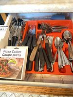 Drawer view showing mixed flatware, knives, tongs, whisks and orange plastic tray