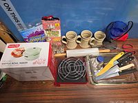 Full lot layout showing boxed waffle maker, steins, utensils, bakeware, blue glass bowl on stand, cleaning supplies