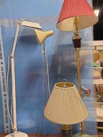 Three lamps shown: white adjustable floor/task lamp, small candlestick table lamp, tall brass and burgundy ceramic lamp with red shade