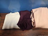 Three folded knit garments: off-white sweater, dark purple dress, blush pink sweater