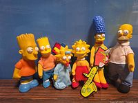 Front view of all five Simpsons plush figures standing together with cardboard accessories visible