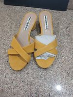 Top view of both yellow suede mule sandals showing criss-cross straps, block heels, brand logo on insoles, and tissue stuffing indicating unused condition