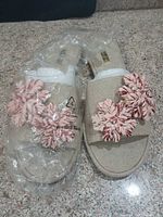 Top view of both sandals showing beige linen material and pink raffia flower accents with protective plastic